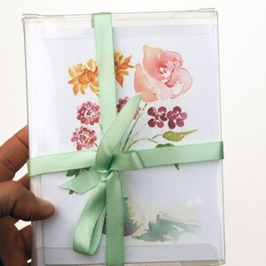 Op de afbeelding: Een set wenskaarten met een aquarel-illustratie van een boeket bloemen in een vaas. De bloemen zijn roze, oranje en paars. De kaarten zijn gebonden met een groen lint.