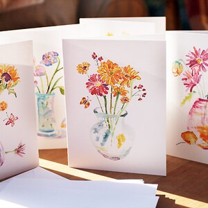 Op de afbeelding: Set van vier aquarel-wenskaarten met bloemstukken in vazen. Elke kaart heeft een ander type bloem, waaronder zonnebloemen, tulpen en gerbera's. De kaarten zijn allemaal wit met een lichtroze rand.