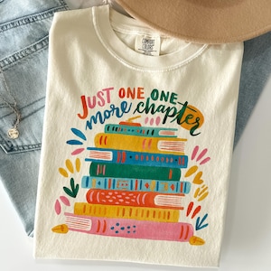 May include: A cream-colored t-shirt featuring a graphic of a stack of colorful books with the text "JUST ONE MORE CHAPTER" in multi-colored lettering. The design includes books in shades of yellow, blue, green, orange, and pink with decorative patterns.