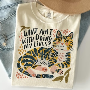 May include: A cream-colored t-shirt featuring a graphic of a reclining tabby cat with dark stripes and pink accents. The text "WHAT AM I DOING WITH MY LIVES?" is printed in black above the cat, surrounded by small orange and pink abstract shapes.