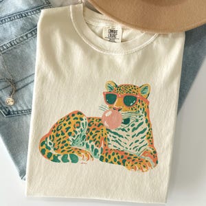 May include: A cream-colored short-sleeve t-shirt featuring a graphic illustration of a leopard wearing red sunglasses and blowing a pink bubblegum bubble. The shirt has a crew neckline and a Comfort Colors brand label printed on the inside collar.