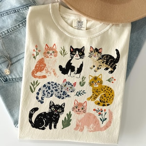 May include: A cream-colored t-shirt featuring a graphic print of seven cats in various colors and patterns, including orange, black and white, tabby, spotted, and pink, interspersed with small green leaf and red berry illustrations.