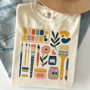 May include: A cream-colored t-shirt featuring a graphic print of various art supplies including a watercolor palette, paintbrushes, jars of paint, a pink flower, and green leaves. The inner neck label displays the text Comfort Colors.
