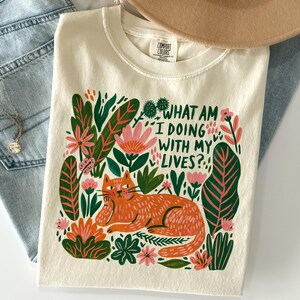 May include: A cream-colored t-shirt featuring a graphic of an orange cat lying among pink flowers and green leaves. The text "WHAT AM I DOING WITH MY LIVES?" is printed in dark green letters above the cat. The shirt has a crew neckline.