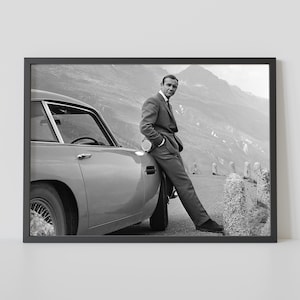 Puede incluir: Una fotografía en blanco y negro de un hombre de pie junto a un coche clásico. El hombre lleva un traje y mira a la cámara. El coche es un coche deportivo vintage con un capó largo y un diseño elegante.