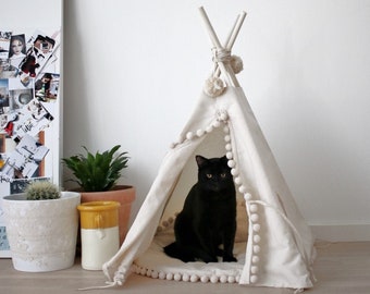 May include: A white teepee-style cat bed with a black cat sitting inside. The bed has a white pom-pom trim and is decorated with white pom-poms.