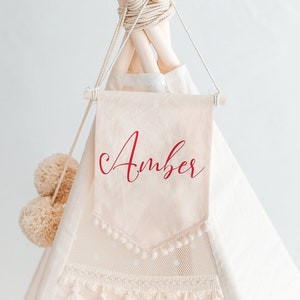 May include: A white teepee tent with a cream-coloured fabric banner that says "Amber" in red cursive lettering. The teepee has a white lace trim and tassels.