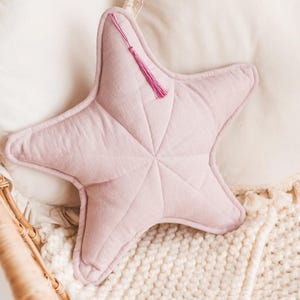 May include: A soft, light pink star-shaped pillow with a decorative tassel, resting on a cream-colored knitted blanket. The pillow is quilted with a starburst pattern. Two round, cream-colored pillows with ruffled edges are in the background.