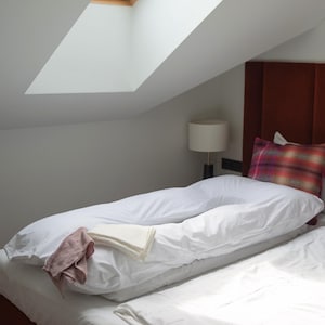 May include: A bed with a white U-shaped pillow, white sheets, and a red headboard. A plaid pillow and a small lamp are also visible. A small window is in the ceiling.