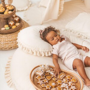 Op de afbeelding: Een jong kind ligt op een wit, pluche bed met een wit kussen en een dienblad met peperkoekjes. De koekjes zijn versierd met glazuur en hebben verschillende vormen, waaronder sterren, harten en kronen. Het kind draagt een wit, mouwloos shirt en heeft bruin haar.