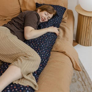 Peut inclure: Un oreiller de corps bleu marine avec un motif floral de petites fleurs rouges, blanches et bleues. L'oreiller est sur un lit avec une literie marron. Une personne est allongée sur l'oreiller, portant un haut marron et un pantalon beige.
