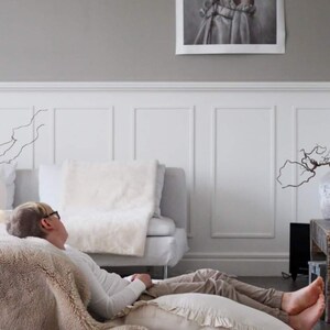 May include: A person wearing a white shirt and glasses is lying on a beige fluffy rug with a white pillow. The person is resting on a large beige throw blanket. The room has white wainscoting and a painting on the wall.