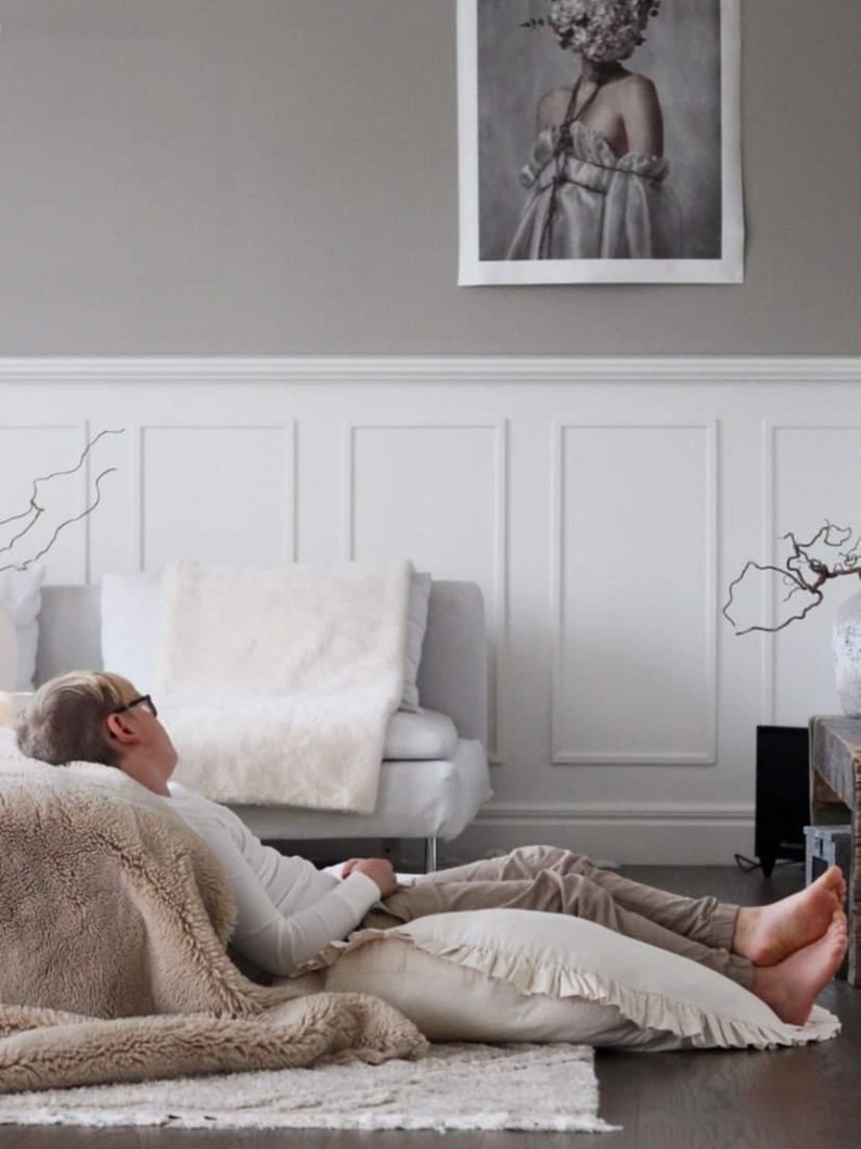 May include: A person wearing a white shirt and glasses is lying on a beige fluffy rug with a white pillow. The person is resting on a large beige throw blanket. The room has white wainscoting and a painting on the wall.
