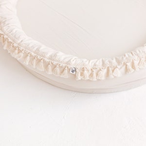 May include: A cream-colored round baby nest with a white tassel trim. The nest is made of soft fabric and has a comfortable, supportive design.