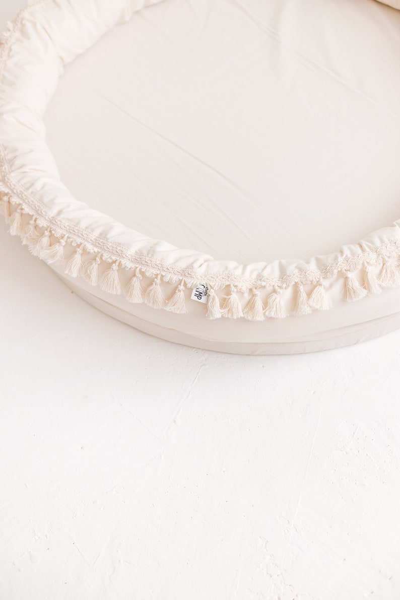 May include: A cream-colored round baby nest with a white tassel trim. The nest is made of soft fabric and has a comfortable, supportive design.