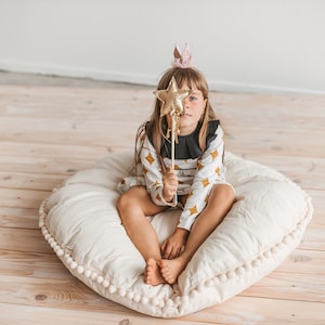 May include: A young girl wearing a white and gold dress with a black collar and a pink crown sits on a large white pillow with a pom-pom trim. She is holding a gold star wand in front of her face.