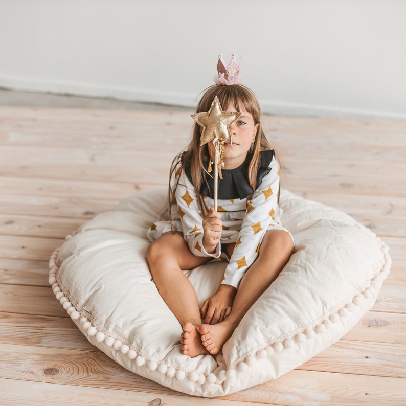 May include: A young girl wearing a white and gold dress with a black collar and a pink crown sits on a large white pillow with a pom-pom trim. She is holding a gold star wand in front of her face.