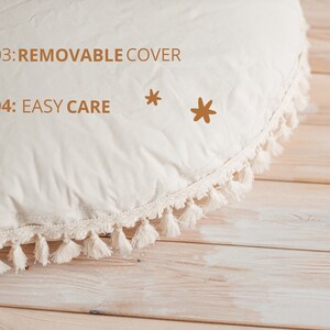 May include: A cream-colored round cushion with a white tassel trim. The cushion has the text "03: REMOVABLE COVER" and "04: EASY CARE" printed on it. Two brown stars are also printed on the cushion.