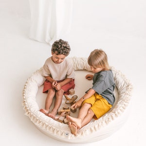 May include: Two children sitting in a round, white, fringed cushion playing with wooden blocks and toys. The cushion has a white background with a white fringe around the edge.
