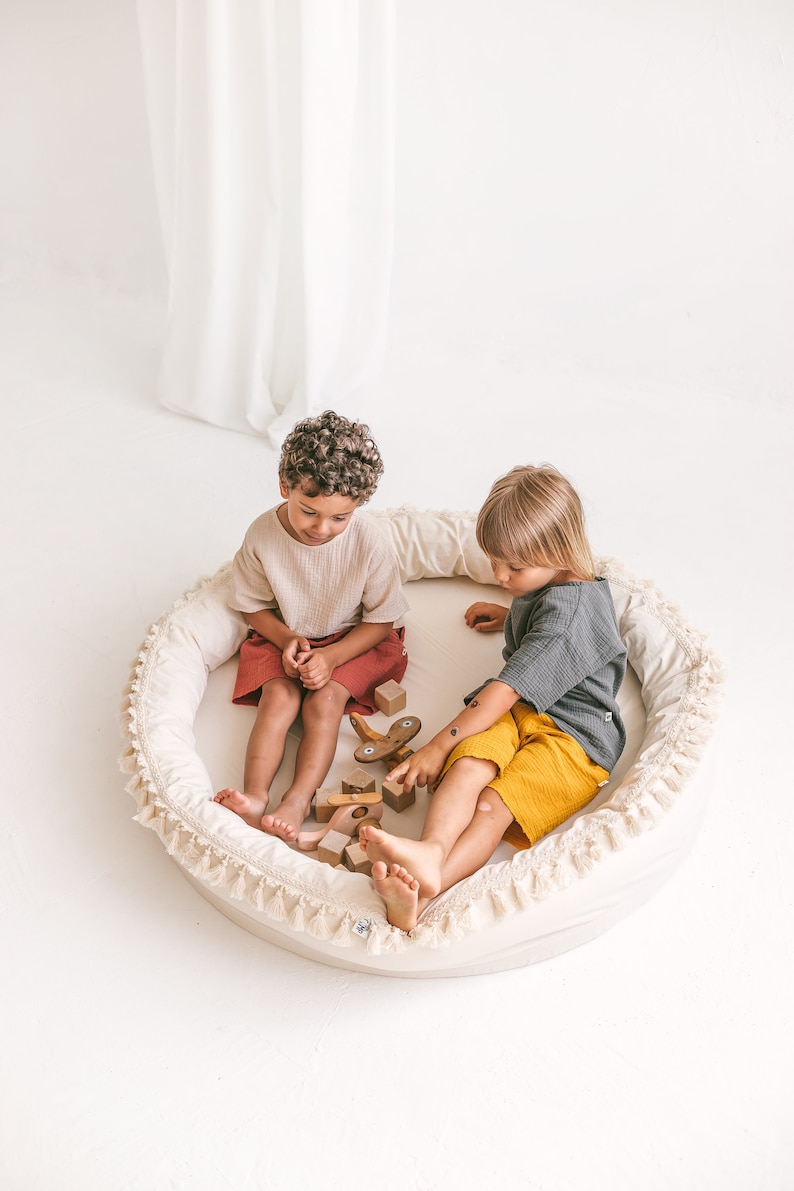 May include: Two children sitting in a round, white, fringed cushion playing with wooden blocks and toys. The cushion has a white background with a white fringe around the edge.