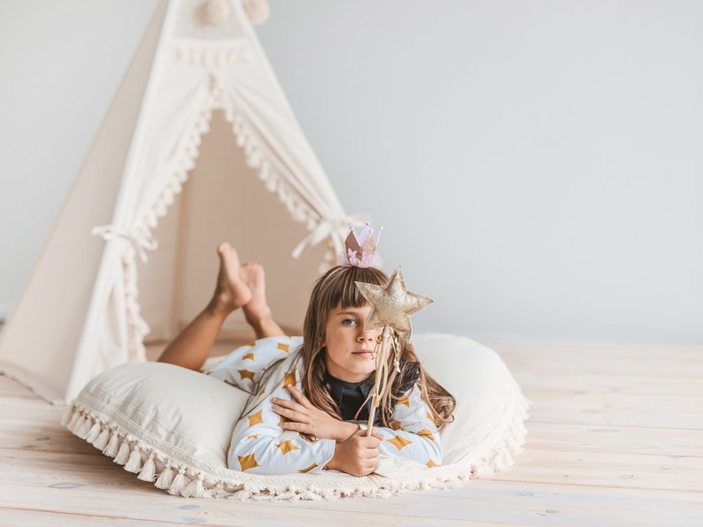 May include: A young girl wearing a crown and pajamas lies on a white pillow with a gold star wand. She is in front of a white teepee tent.