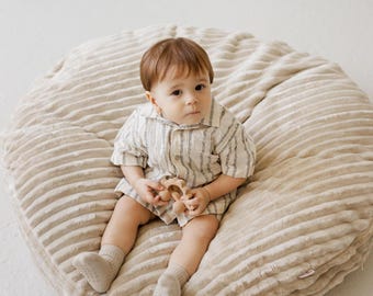 May include: A baby wearing a striped shirt and shorts, holding a wooden toy, sits on a large, beige, corduroy-textured beanbag. The baby is also wearing beige socks. The beanbag is on a white surface.