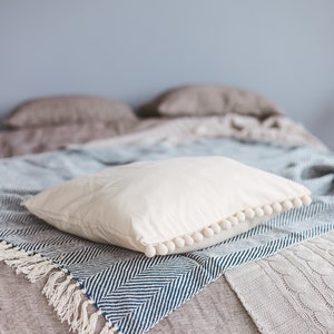 May include: A white pillow with a pom-pom trim sits on a blue and white striped throw blanket with fringe. The blanket is on top of a gray linen bedspread.