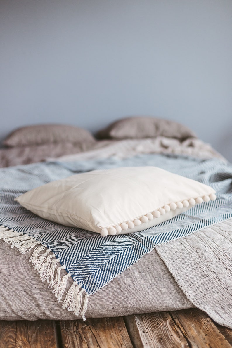 May include: A white pillow with a pom-pom trim sits on a blue and white striped throw blanket with fringe. The blanket is on top of a gray linen bedspread.