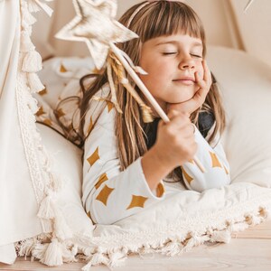 May include: A young girl with long brown hair is lying in a white teepee with a gold star-shaped wand. She is wearing a white and gold patterned shirt and has her eyes closed.