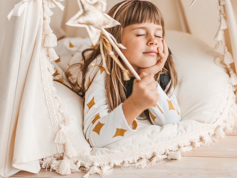 May include: A young girl with long brown hair is lying in a white teepee with a gold star-shaped wand. She is wearing a white and gold patterned shirt and has her eyes closed.