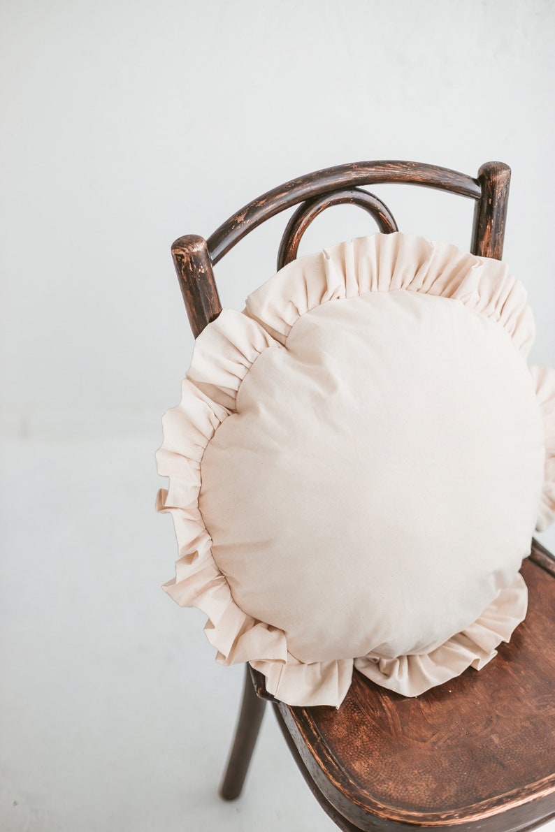 May include: A round, cream-colored pillow with a ruffled edge sits on a wooden chair. The chair has a dark brown finish and a curved back.