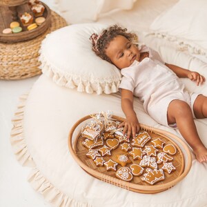 May include: A child lays on a cream-colored round rug with a tray of gingerbread cookies in front of them. The cookies are decorated with icing and have various shapes, including stars, hearts, and crowns. The tray is made of woven wicker and has a light brown color.