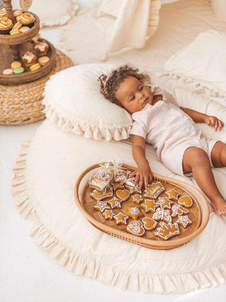 May include: A child lays on a cream-colored round rug with a tray of gingerbread cookies in front of them. The cookies are decorated with icing and have various shapes, including stars, hearts, and crowns. The tray is made of woven wicker and has a light brown color.