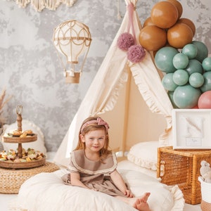 May include: A young girl wearing a brown dress with a lace collar sits on a large, white, round pillow with ruffles.  She is smiling and looking at the camera.  Behind her is a teepee tent decorated with pink pom-poms and a hot air balloon decoration.  There are also brown and green balloons in the background.