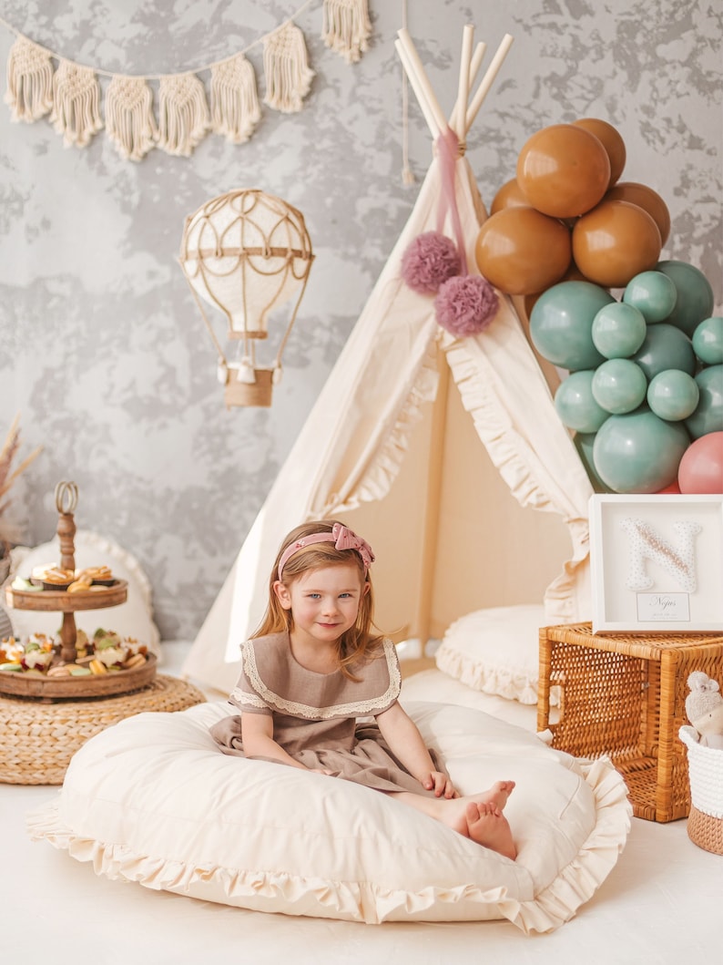 May include: A young girl wearing a brown dress with a lace collar sits on a large, white, round pillow with ruffles.  She is smiling and looking at the camera.  Behind her is a teepee tent decorated with pink pom-poms and a hot air balloon decoration.  There are also brown and green balloons in the background.