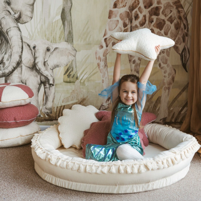 May include: A young girl wearing a blue and green dress sits in a cream-colored, round, cushioned bed with a white fringe. She is holding a white star-shaped pillow above her head. The bed is in front of a wall with a mural of elephants and giraffes.