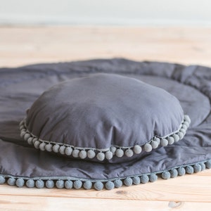 May include: A gray round pillow with a gray pom-pom trim sits on a gray round play mat with a gray pom-pom trim.
