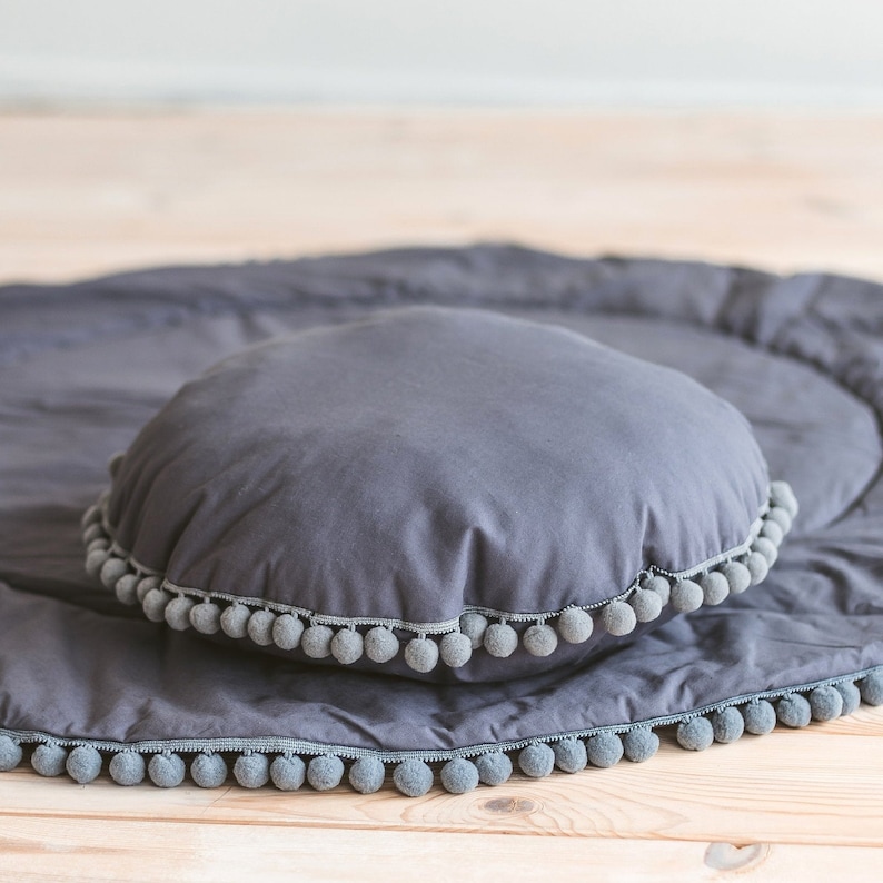 May include: A gray round pillow with a gray pom-pom trim sits on a gray round play mat with a gray pom-pom trim.