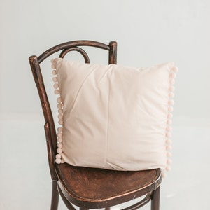 May include: A light pink pillow with a pom-pom trim sits on a dark brown wooden chair with a curved back.