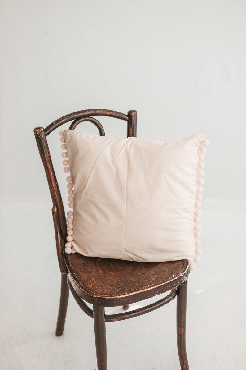 May include: A light pink pillow with a pom-pom trim sits on a dark brown wooden chair with a curved back.