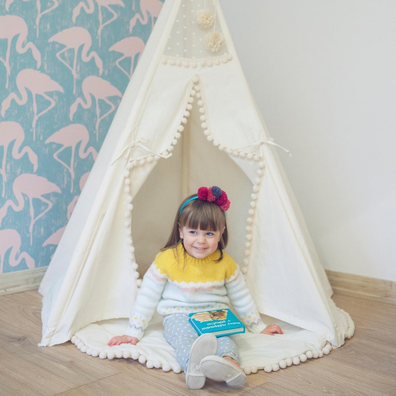 May include: A young girl wearing a yellow and pink sweater sits inside a white teepee tent with a pom-pom trim. She is holding a blue book with the title "Огромный мир" in Cyrillic script. The teepee is set up in a room with a pink flamingo wallpaper.