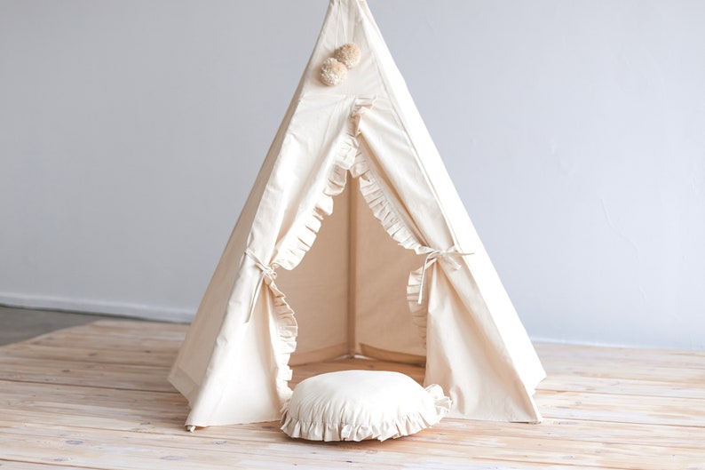 May include: A white canvas teepee tent with a ruffled edge and pom poms on top. The teepee is set up on a wooden floor and has a white pillow inside.