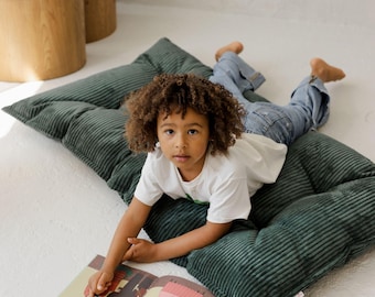 Puede incluir: Un niño está acostado sobre un cojín de suelo de pana verde oscuro, leyendo un libro con ilustraciones coloridas. El cojín es rectangular y tiene una textura acanalada. El niño lleva una camiseta blanca y vaqueros azules.