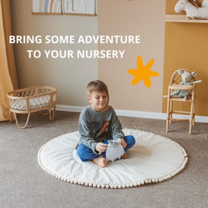 May include: A young child sits on a white play mat with a pom-pom trim, holding a gray stuffed animal. The mat is on a beige carpet. The child is wearing a gray long-sleeved shirt and blue pants. The background features a brown wall with a yellow star sticker and a wooden shelf with a stuffed animal on it.