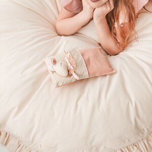 May include: A cream-colored round play mat with a ruffled edge. A small stuffed mouse with a pink dress is lying on a striped pillow on the mat.