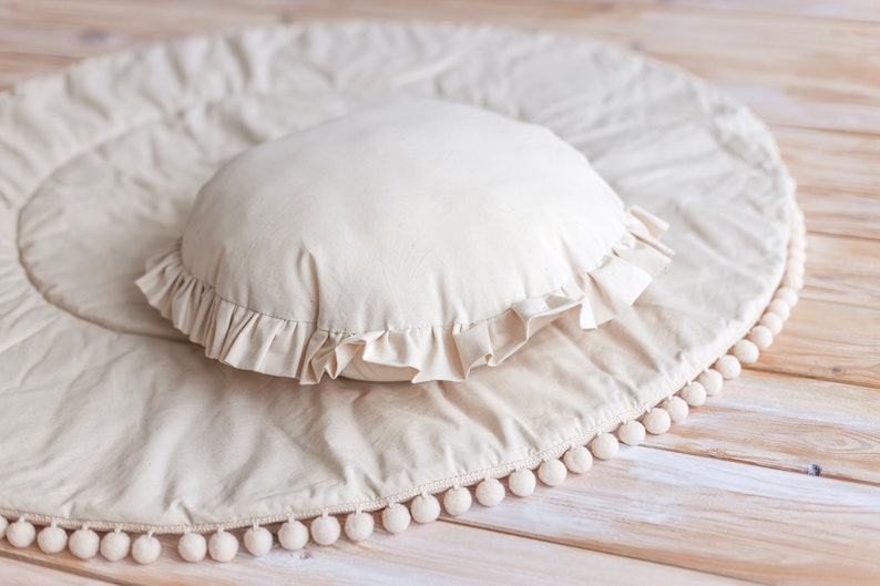 May include: A cream-colored round pillow with a ruffled edge sits on a cream-colored round play mat with a white pom-pom trim.