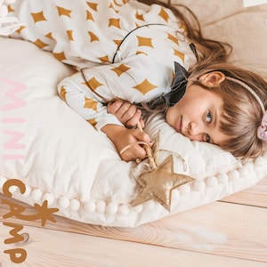 May include: A white and gold star-shaped toy wand with a white pom-pom trim pillow. The pillow has the words "mini camp" written on it in pink and brown letters. A young girl is lying on the pillow with her head resting on the pillow. She is wearing a white and gold patterned dress with a black bow.