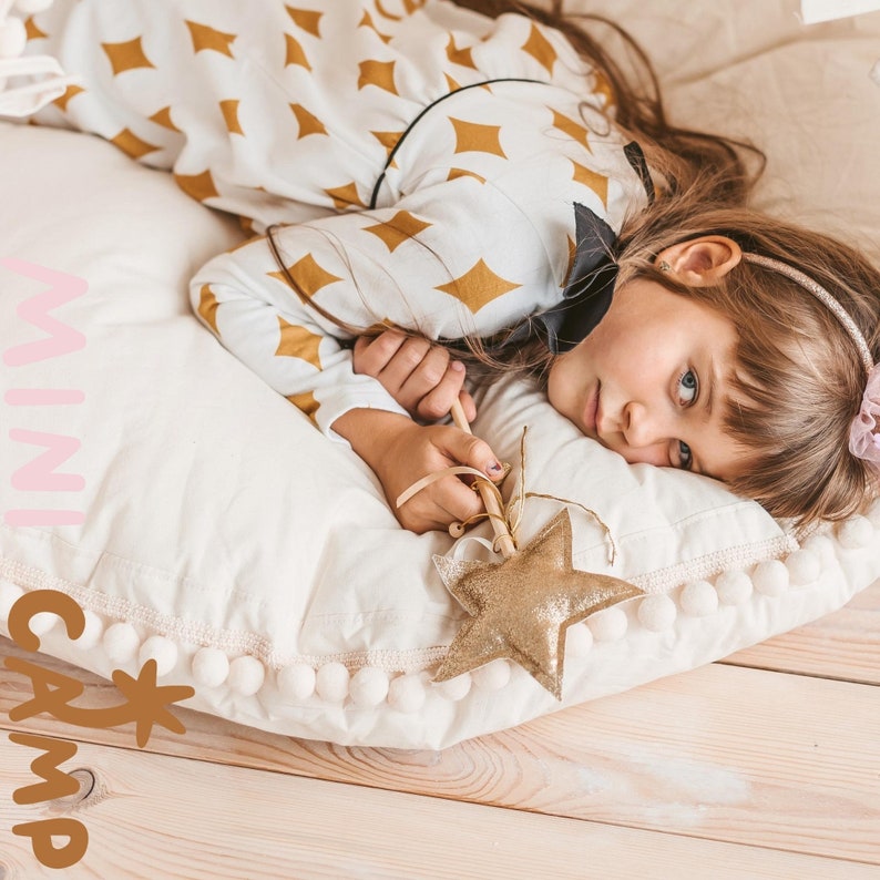 May include: A white and gold star-shaped toy wand with a white pom-pom trim pillow. The pillow has the words "mini camp" written on it in pink and brown letters. A young girl is lying on the pillow with her head resting on the pillow. She is wearing a white and gold patterned dress with a black bow.