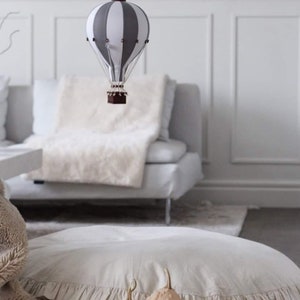 May include: A white and gray striped hot air balloon hanging from the ceiling above a white couch with a fluffy white throw blanket. Two small beige gourds sit on a white ruffled floor pillow in front of the couch.