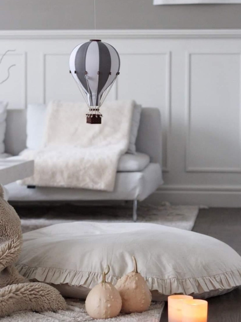 May include: A white and gray striped hot air balloon hanging from the ceiling above a white couch with a fluffy white throw blanket. Two small beige gourds sit on a white ruffled floor pillow in front of the couch.